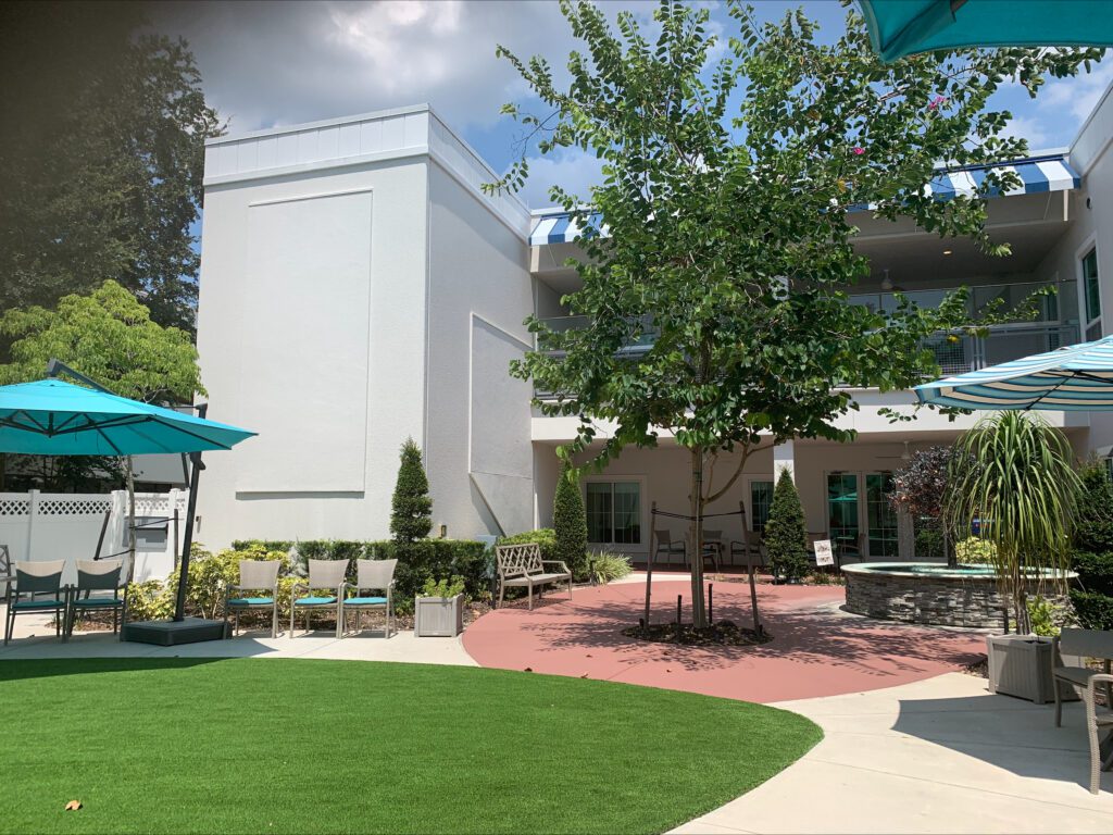 Image of a senior living community's outdoor courtyard with plenty of umbrella covered seating, garden boxes, and sunshine.
