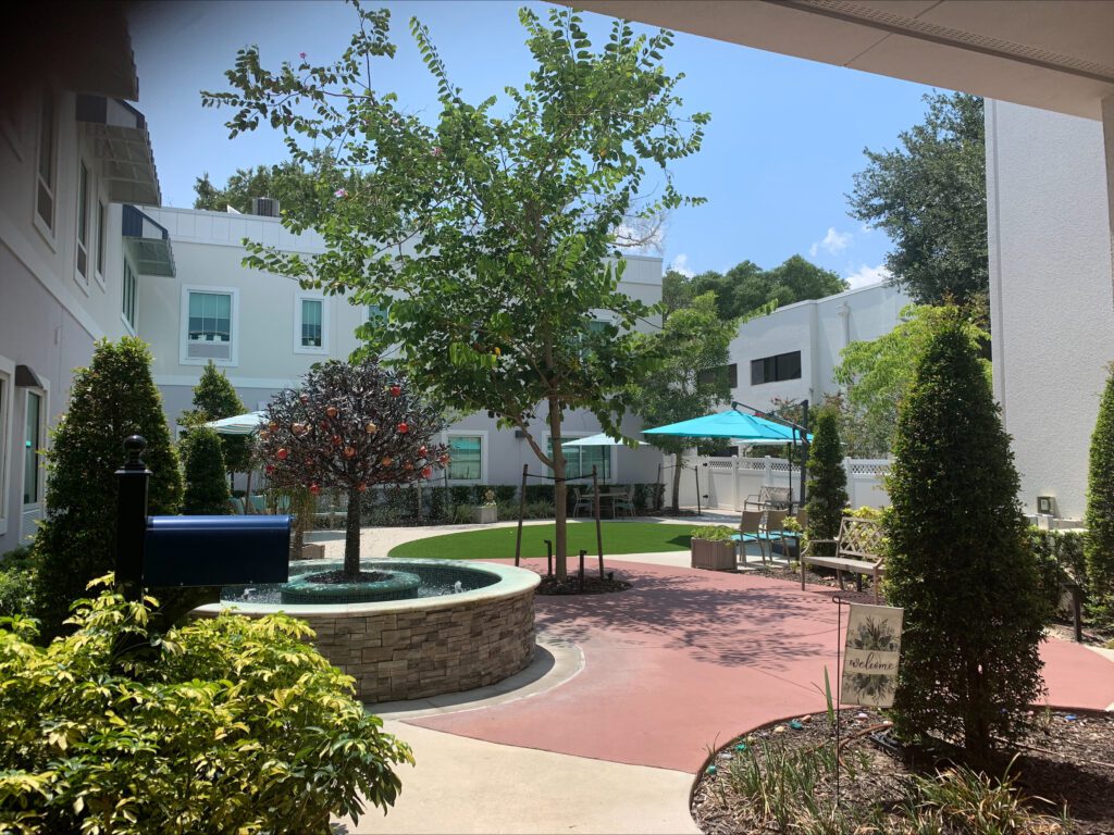 Image of a senior living community's outdoor courtyard with plenty of umbrella covered seating, garden boxes, and sunshine.